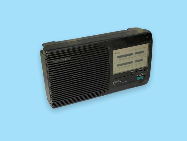 Może przedstawiać: Czarno-białe radio Sony FM/AM w stylu vintage z wyraźnym wyświetlaczem pokazującym częstotliwości radiowe. Radio ma prostokątny kształt z żebrowaną osłoną głośnika i selektorem pasma. Radio znajduje się na jasnoniebieskim tle.