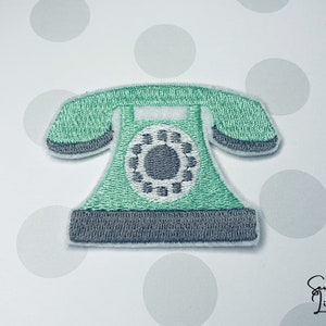 Pode incluir: Um remendo bordado em forma de telefone de disco verde com um dial cinza e detalhes brancos. O remendo tem uma borda branca.