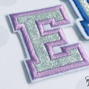 May include: A silver glitter letter E with a purple outline. The letter is embroidered and has a raised, textured look.