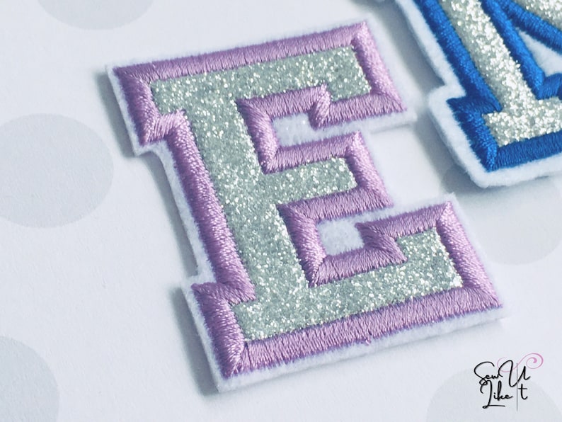 May include: A silver glitter letter E with a purple outline. The letter is embroidered and has a raised, textured look.