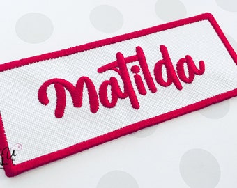 May include: A white rectangular patch with a pink border and the name "Matilda" embroidered in pink.