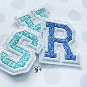 May include: Three felt letter patches in blue and turquoise with white stitching. The letters are "S", "R", and "T".