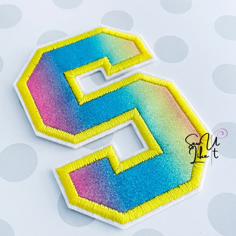 May include: A rainbow ombre felt letter S with a yellow satin stitch outline. The letter is on a white background with gray polka dots.