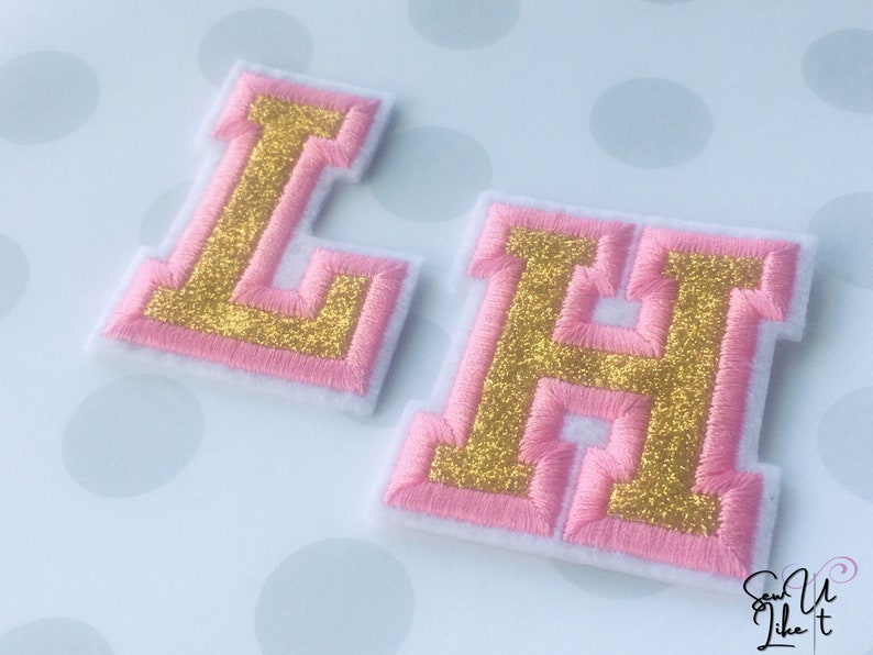 May include: Two felt letters, L and H, with pink outlines and gold glitter inside. The letters are on a white background with pink polka dots.