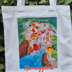 May include: White canvas tote bag featuring a colourful illustration of a town on a cliffside with the text "Riomaggiore" printed in red.