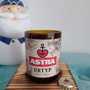 Könnte beinhalten: Ein braunes Glas-Kerzengefäß mit einem Etikett, auf dem "ASTRA URTYP SEIT 1909" steht. Das Etikett zeigt ein rotes Herz mit einem Anker darin und ein Totenkopf- und Knochen-Design. Das Kerzengefäß steht auf einem Holz-Untersetzer mit einem Flaschenverschluss, auf dem "URTYP" steht.