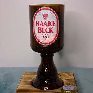 Op de afbeelding: Een uniek glas gemaakt van een bruine bierfles, met het label "HAAKE BECK Pils". Het staat op een houten voet, met een flesdop ernaast. Een creatief upcycle-item.