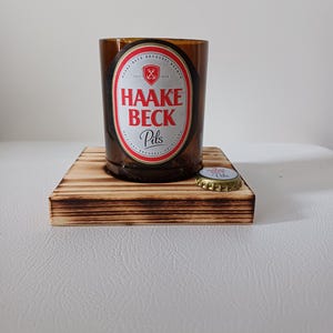 Può includere: Un bicchiere marrone riciclato con l'etichetta "HAAKE BECK Pils", posto su una piccola base quadrata di legno bruciato. Un tappo di bottiglia è appoggiato accanto. Un oggetto unico e riciclato.