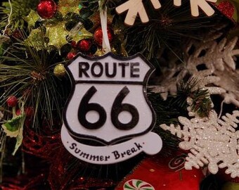Op de afbeelding: Kerstboomversiering met een Route 66-bord met de tekst "Summer Break". De versiering is wit met zwarte letters en is omgeven door feestelijke decoraties, waaronder rood lint en sneeuwvlokken.