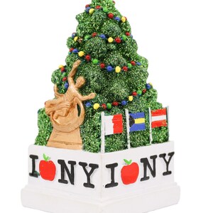 Può includere: Un ornamento per albero di Natale verde glitterato con una statua dorata di una persona che tiene una torcia, circondata da bandiere. La base dell'ornamento è bianca con il testo "I  NY I  NY" e mele rosse.