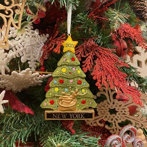 Può includere: Un ornamento per albero di Natale a forma di albero di Natale verde con glitter, con una stella dorata in cima e una base dorata. L'ornamento ha un nastro rosso e bianco a righe e il testo "2021" sulla stella. La base dell'ornamento ha il testo "NEW YORK" su di essa.