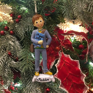 May include: A blue and white Christmas ornament depicting a boy holding a book that says "5th grade 2020". The ornament has a white base with the name "Maddox" written on it.