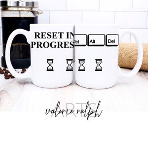 May include: Two white ceramic mugs with black text and graphics. The text on the mugs reads "Reset in Progress" with computer keys labeled "Ctrl", "Alt", and "Del" and four hourglass icons.