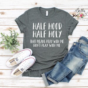 Peut inclure: Un t-shirt gris avec le texte "HALF HOOD HALF HOLY THAT MEANS PRAY WITH ME DON'T PLAY WITH ME". L'image comprend un jean bleu et des baskets blanches Converse. Une petite plante est &eacute;galement pr&eacute;sente.