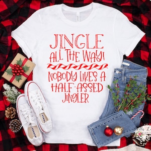 May include: White t-shirt with red text that reads "Jingle All The Way! Nobody Likes A Half-Assed Jingler". The shirt is laid out on a red and black plaid fabric with a pair of white sneakers, blue jeans, and a small Christmas-themed gift box.