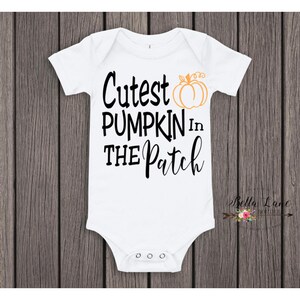 May include: White baby onesie with the text "Cutest PUMPKIN In THE Patch" in black script. An orange pumpkin graphic is above the word "Patch". The onesie has short sleeves and snaps at the bottom. The background is a wooden surface.