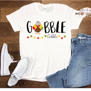 May include: White t-shirt with a black graphic that says "Gobble" with a cartoon turkey in the middle of the word. The graphic also includes a row of colorful dots below the word.