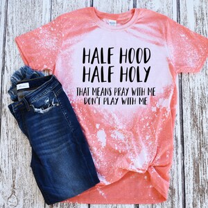 Peut inclure: Un t-shirt corail avec des taches de javel blanches et le texte "HALF HOOD HALF HOLY THAT MEANS PRAY WITH ME DON'T PLAY WITH ME" en noir. Un jean bleu fonc&eacute; est pli&eacute; &agrave; c&ocirc;t&eacute; du t-shirt.