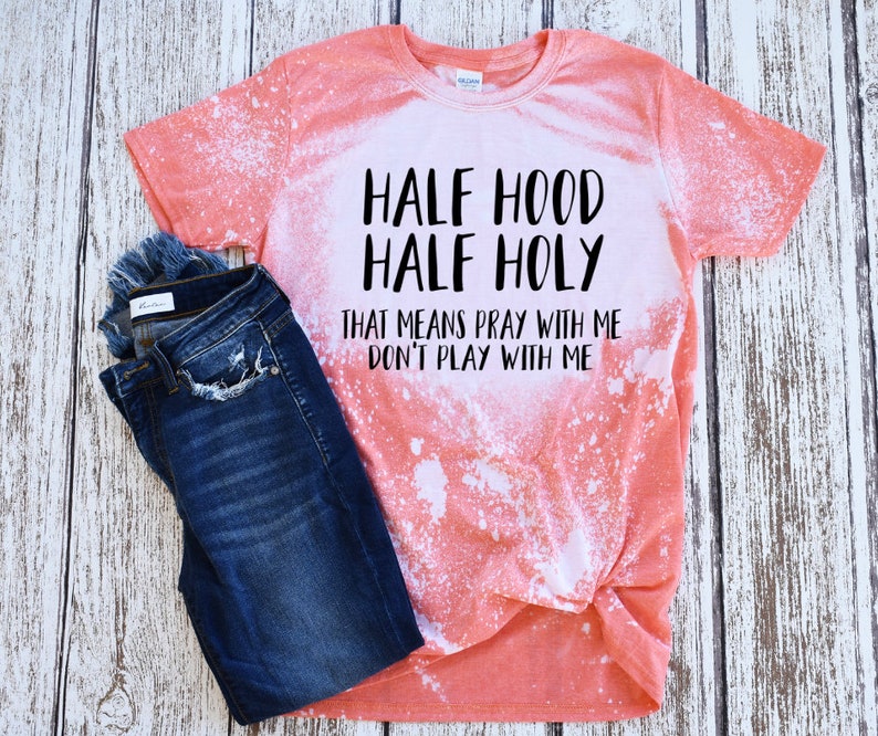 Peut inclure: Un t-shirt corail avec des taches de javel blanches et le texte "HALF HOOD HALF HOLY THAT MEANS PRAY WITH ME DON'T PLAY WITH ME" en noir. Un jean bleu fonc&eacute; est pli&eacute; &agrave; c&ocirc;t&eacute; du t-shirt.