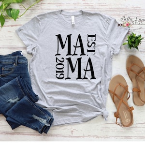 May include: A light gray t-shirt with the word "MAMA" in large black letters, with "EST. 2019" printed vertically on the left side. The shirt is paired with blue jeans and brown sandals.