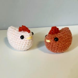 May include: Two small, round, crocheted chicken figures. One chicken is light cream-colored, and the other is a medium brown. Both feature a bright red crocheted comb on top, small yellow beaks, and round, black plastic bead eyes.