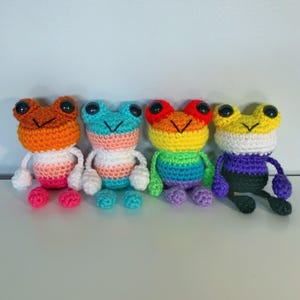 Peut inclure: Quatre peluches grenouilles en crochet faites à la main, alignées. Chaque grenouille possède de grands yeux en plastique noir et une bouche cousue en forme de V. Elles présentent des motifs de couleurs horizontaux distincts : orange et rose, bleu clair et blanc, arc-en-ciel, et jaune, blanc, violet et noir.