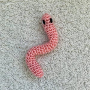 May include: A handmade, pink crocheted worm with black eyes. The worm is made of a soft, textured yarn and is curled in a gentle curve. The background is a light, textured surface.