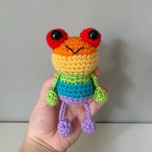 May include: A colourful, handmade crochet frog. The frog features a rainbow design with red eyes, a yellow body, and a striped belly in green, blue, and purple. It has green arms and purple legs. A whimsical, handcrafted item, perfect as a gift or decoration.