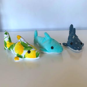 May include: Three plush shark toys. One shark is covered in a lemon print, another is light blue, and the third is dark blue with white spots. Each shark has black button eyes and white accents.