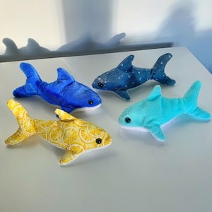 May include: Four plush shark toys in different patterns and colors. One is bright blue, one is turquoise, one is dark blue with a white star pattern, and one is yellow with a repeating lemon slice print. Each shark has black plastic eyes.