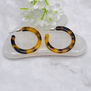 May include: A pair of tortoiseshell hoop earrings. The earrings are a warm brown and black colour, with a classic circular design. The earrings are displayed on a white surface with white flowers in the background.