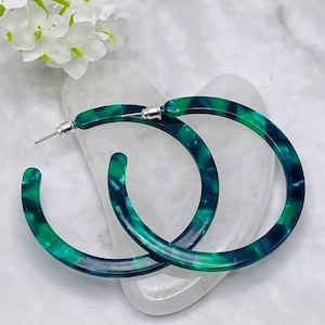 May include: A pair of large hoop earrings with a marbled green and blue pattern. The earrings have a post closure. The earrings are a fashion accessory. The earrings are set against a white background.