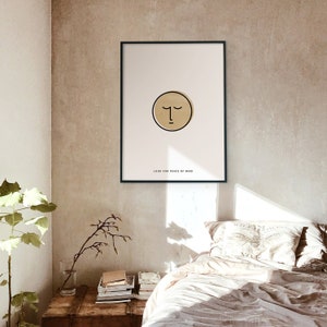 Könnte beinhalten: Gerahmter Kunstdruck mit minimalistischem Gesichtsdesign in einem goldenen Kreis auf cremefarbenem Hintergrund. Der Text "LOOK FOR PEACE OF MIND" steht darunter. Der Rahmen ist schwarz. Das Bild hängt an einer Wand.