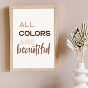 May include: A framed print with the text "ALL COLORS ARE beautiful" in brown, peach, and white lettering on a white background. The frame is light brown wood. The print is hanging on a light brown wall.