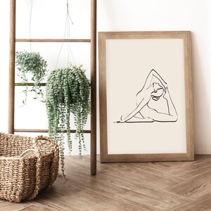 May include: A framed print of a line drawing of a person in a yoga pose. The print is on a light beige background and is framed in a natural wood frame. The print is hanging on a white wall.