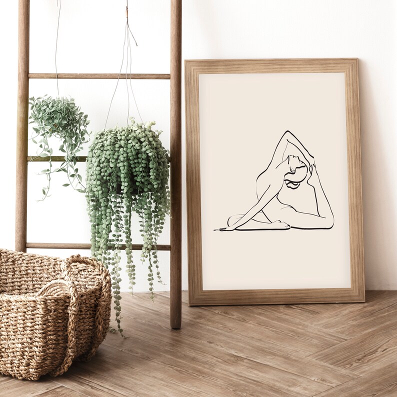 May include: A framed print of a line drawing of a person in a yoga pose. The print is on a light beige background and is framed in a natural wood frame. The print is hanging on a white wall.