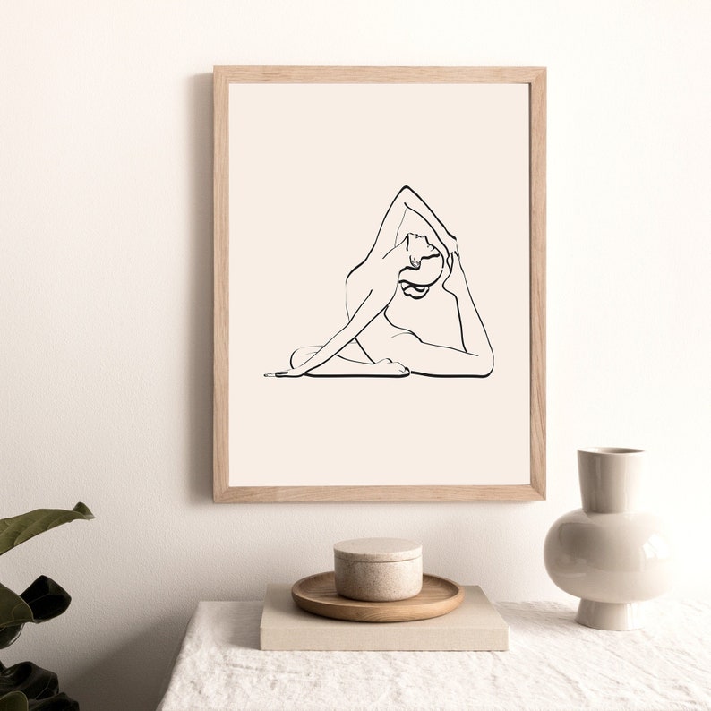 May include: A black and white line drawing of a person in a yoga pose, framed in a light wood frame. The print is on a white background and is displayed on a table with a vase and a small container.