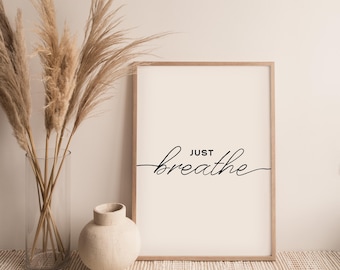 May include: A framed print with a white background and black text that reads "JUST breathe". The print is hanging on a wall with a vase of dried pampas grass in front of it.