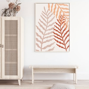 May include: A minimalist wall art print featuring two stylized fern leaves in shades of peach, orange, and terracotta against a white background. The print is framed in a thin, white frame.