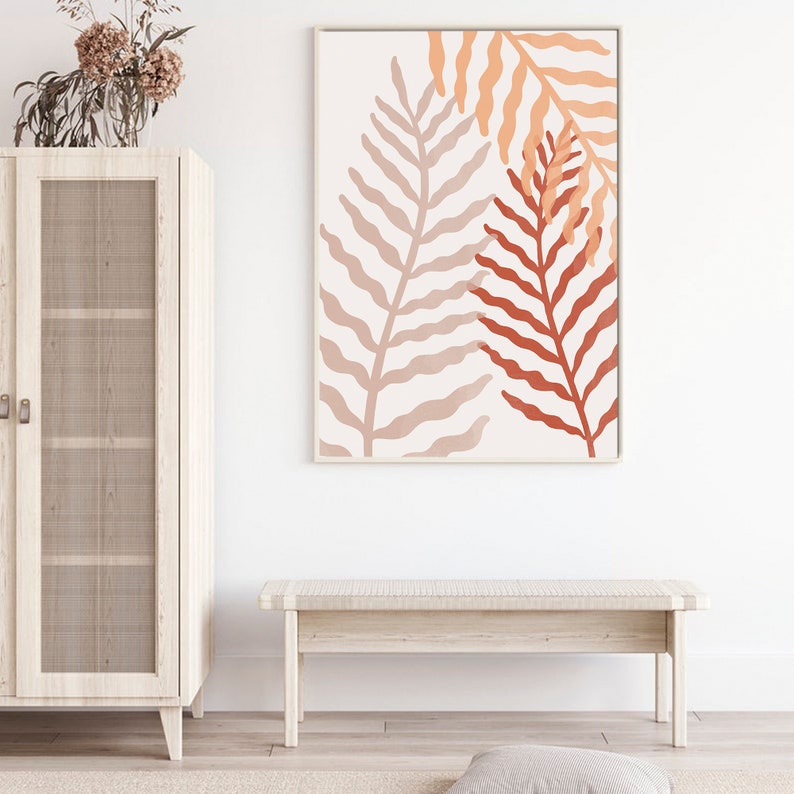 May include: A minimalist wall art print featuring two stylized fern leaves in shades of peach, orange, and terracotta against a white background. The print is framed in a thin, white frame.