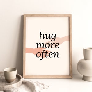Puede incluir: Una impresi&oacute;n enmarcada con un fondo melocot&oacute;n y dos manos que se acercan la una a la otra. El texto "hug more often" est&aacute; impreso en negro.
