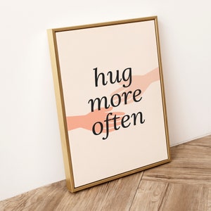 Puede incluir: Una impresi&oacute;n en lienzo enmarcada en dorado con un fondo melocot&oacute;n y dos manos rosas que se acercan la una a la otra. El texto "hug more often" est&aacute; impreso en negro.