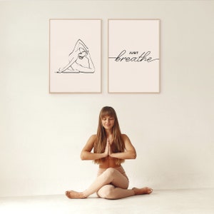 May include: Two framed prints on a white wall. The print on the left shows a line drawing of a person in a yoga pose. The print on the right has the text "JUST breathe" in a cursive font. A woman is sitting in a yoga pose in front of the prints.