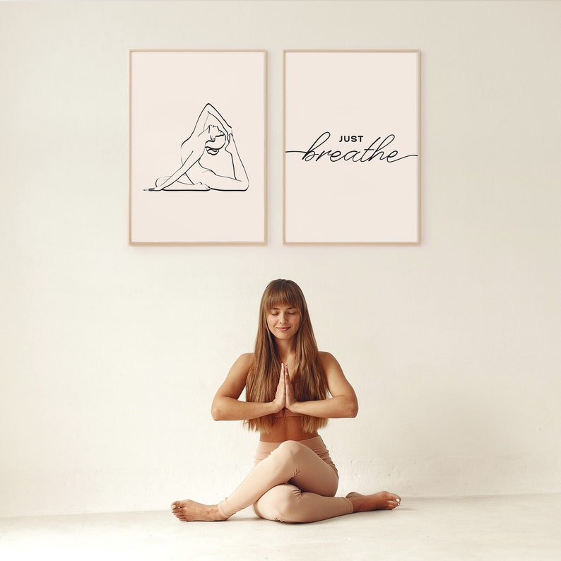 May include: Two framed prints on a white wall. The print on the left shows a line drawing of a person in a yoga pose. The print on the right has the text "JUST breathe" in a cursive font. A woman is sitting in a yoga pose in front of the prints.