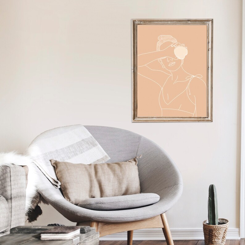 May include: A line drawing of a woman in a swimsuit holding a peach in front of her face. The drawing is on a peach background and is framed in a light wood frame.