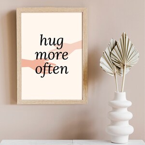 Puede incluir: Una impresi&oacute;n enmarcada con fondo blanco y el texto "hug more often" en negro. Dos manos rosas se acercan la una a la otra en el centro de la impresi&oacute;n.