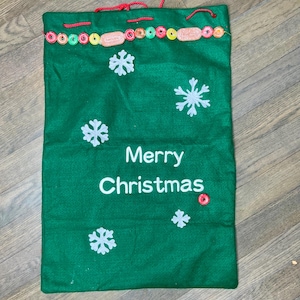Puede incluir: Bolsa de regalo navideña verde con copos de nieve blancos y la frase "Merry Christmas". La bolsa tiene un cordón rojo y está decorada con adornos circulares y rectangulares de colores a lo largo del borde superior.