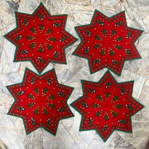 Può includere: Quattro tovagliette rosse a forma di stella con bordo verde. Ogni tovaglietta presenta un motivo ripetuto di alberi di Natale e figure verdi. Le tovagliette sono disposte su una superficie chiara e strutturata.