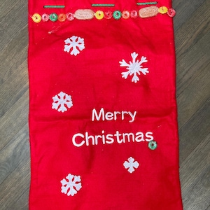 Puede incluir: Bolsa de regalo navideña roja con copos de nieve blancos y la frase "Merry Christmas". La parte superior de la bolsa está decorada con caramelos de colores y un cordón verde. Un artículo festivo.