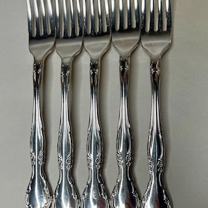 May include: A set of five silver-toned metal forks arranged side by side. Each fork features a polished finish and a decorative handle with embossed floral detailing at the base and along the stem, ending in a rounded, ornate tip.